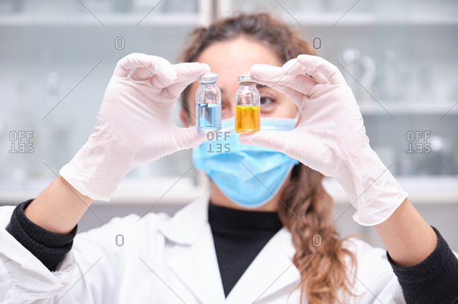 Young female scientist wearing face mask holding two coronavirus vaccine blue and orange vials. Covid-19 vaccine development.