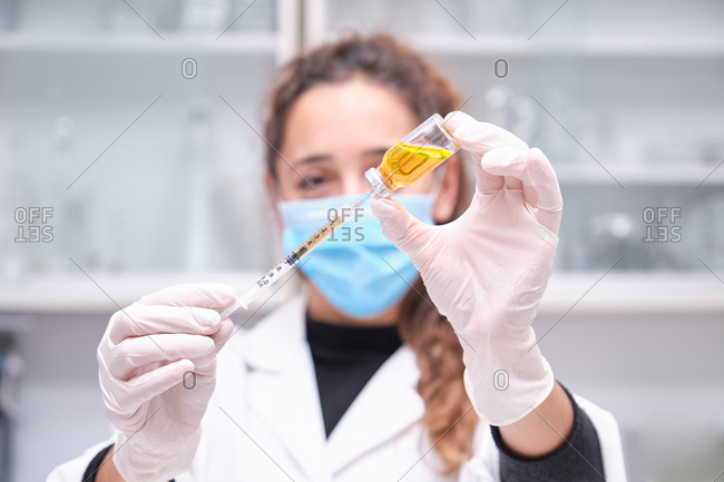 Young female scientist wearing face mask holding a coronavirus vaccine vial, syringe and needle. Covid-19 vaccine development.