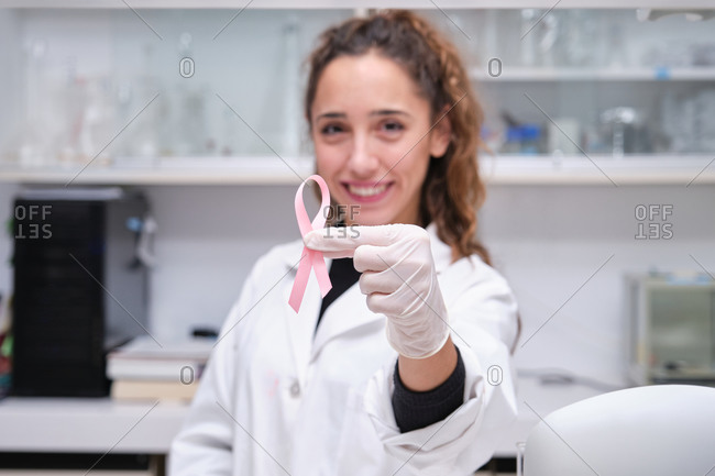 Young female scientist holding a pink ribbon in front of her face, for the breast cancer awareness. Cancer research concept.