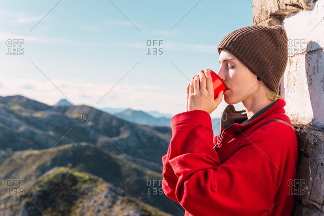 Side view of androgynous traveler standing in highlands on sunny day and enjoying hot drink during trip in El Mazuco with eyes closed