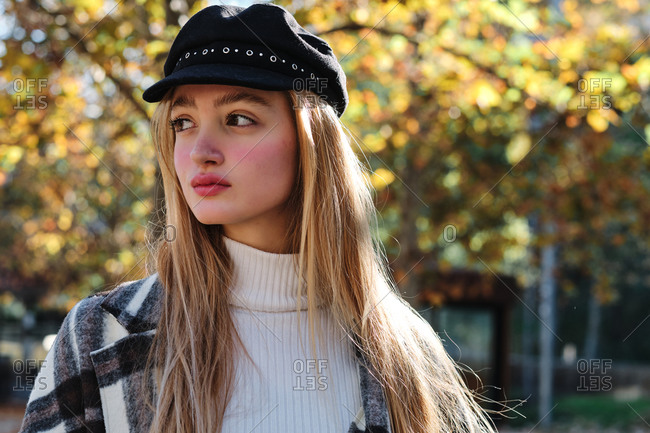 Tranquil young long haired blonde female in stylish hat and checkered coat enjoying sunny weather standing looking away in autumn park