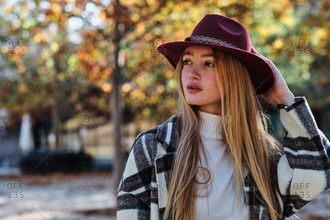 Tranquil young long haired blonde female in stylish hat and checkered coat enjoying sunny weather standing looking away in autumn park