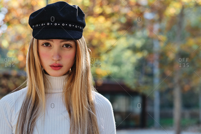 Attractive teen blonde female in turtleneck sweater and stylish cap looking away while standing against blurred background of autumn trees in park