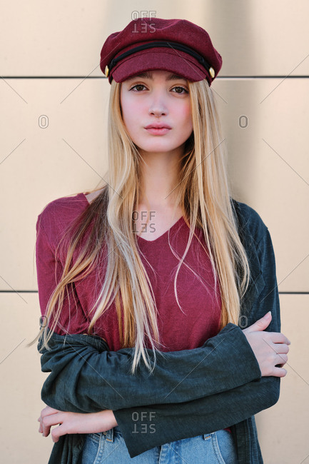 Stylish modern young blonde female in trendy autumn outfit with red cap and coat standing in sunlight against beige wall looking at camera