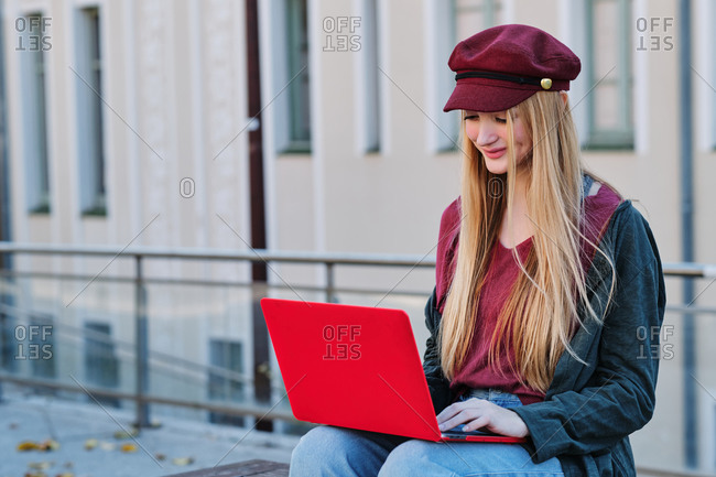 Positive millennial female student in trendy autumn outfit and hat browsing red laptop while sitting on paved terrace on city street