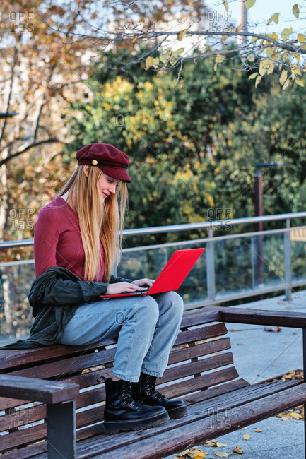 Full body of focused millennial female in trendy casual clothes and cap sitting on wooden bench and browsing modern red laptop while spending autumn day in park