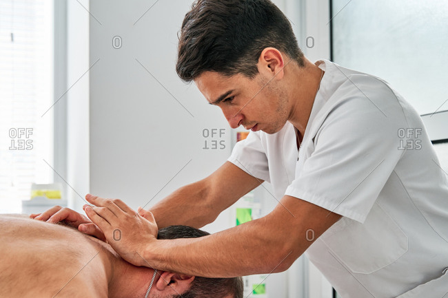 Side view of osteopath treating neck of male patient in mask lying on table in medical room during rehabilitation procedure