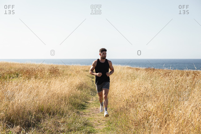 Muscular male jogger running along path in meadow during training on background of sea in summer