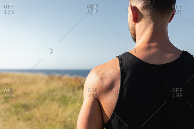Determined male athlete with strong torso standing on grassy hill on sunny day in summer and looking away