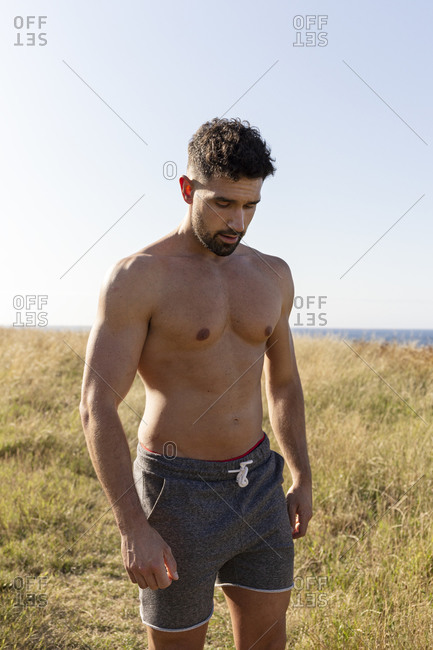 Determined male athlete with strong naked torso standing on grassy hill on sunny day in summer and looking down