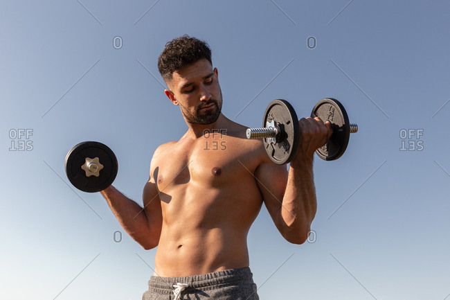 Low angle of handsome male athlete with naked torso doing exercises with dumbbells while standing against blue sky in summer