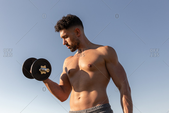 Low angle of handsome male athlete with naked torso doing exercises with dumbbells while standing against blue sky in summer