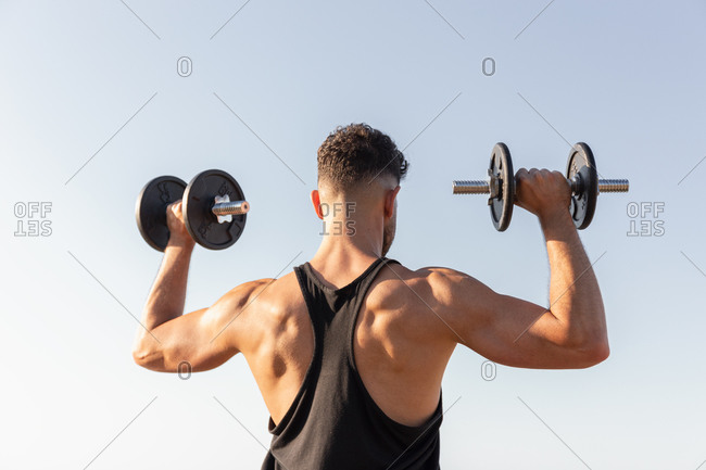 Back view of handsome male athlete with strong torso doing exercises with dumbbells while standing against blue sky in summer