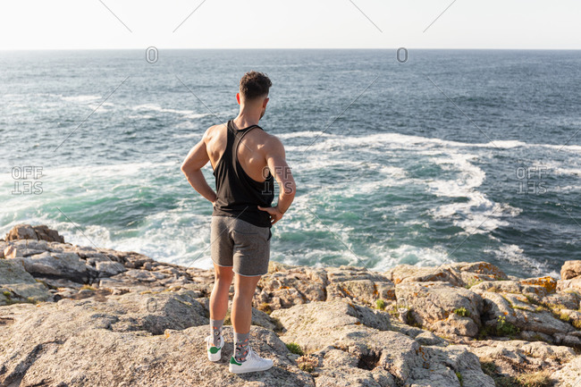 Back view of muscular male athlete in sportswear standing on beach and enjoying sundown after training in summer