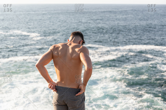 Back view of muscular male athlete with naked torso standing on beach and enjoying sundown after training in summer