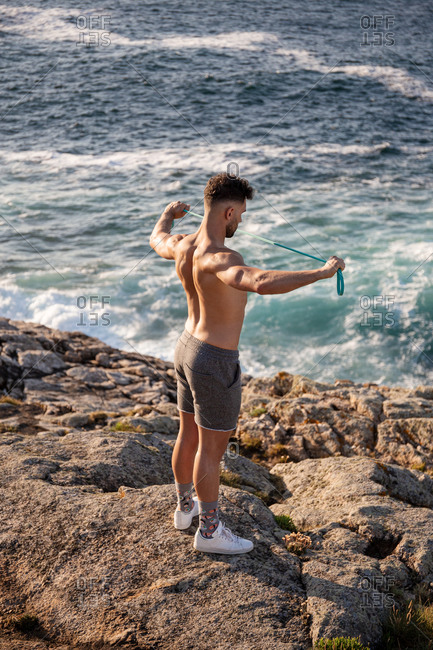 Muscular male bodybuilder with naked torso standing on seashore and doing exercises with resistance band during workout in summer