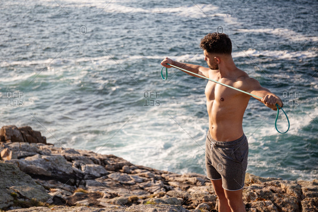 Muscular male bodybuilder with naked torso standing on seashore and doing exercises with resistance band during workout in summer