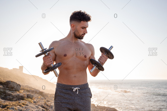 Handsome male athlete with naked torso doing exercises with dumbbells while standing against blue sky in summer