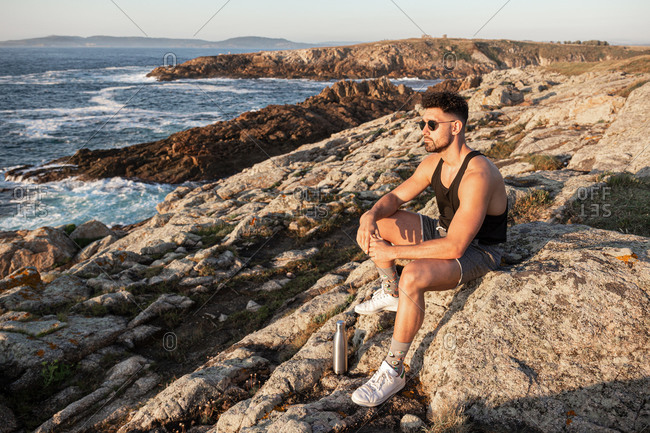 Side view of peaceful male sitting on rock on coast and admiring calm seascape at sundown in summer