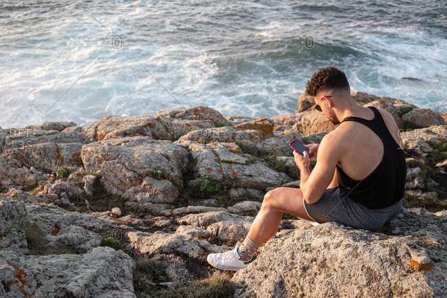 Side view high angle of male in summer clothes sitting on rock near sea and messaging on social media via cellphone at sunset