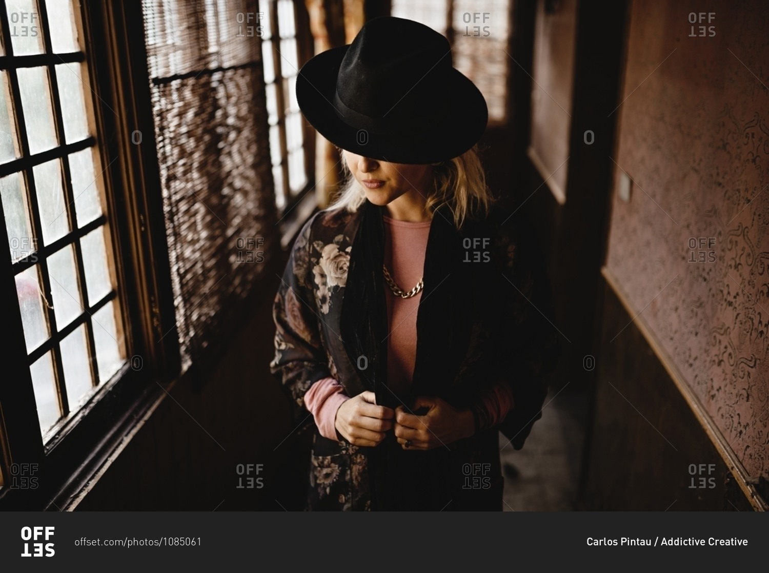 Stylish female in elegant vintage outfit and hat standing in narrow hallway of old house and looking at mirror