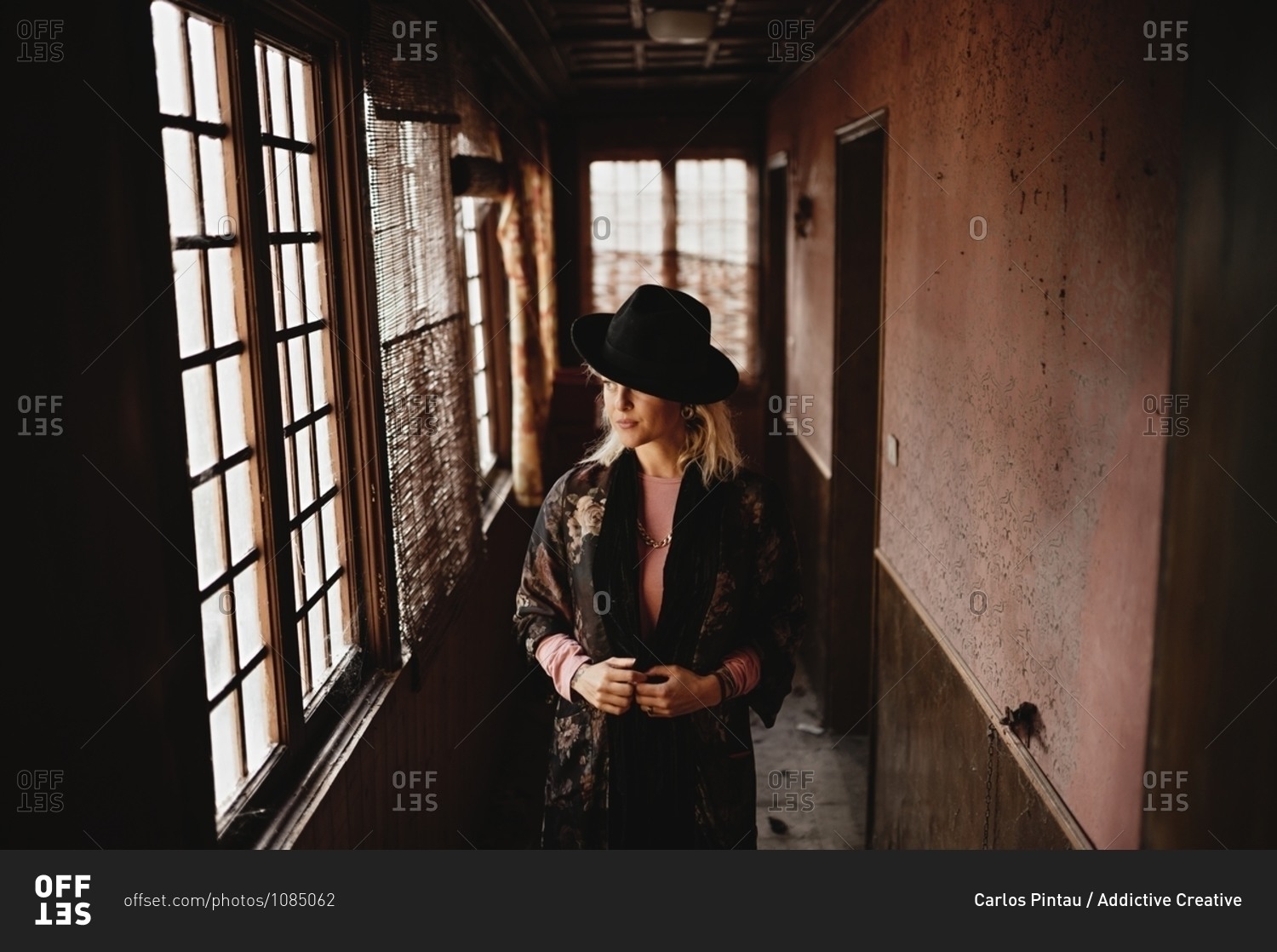 Stylish female in elegant vintage outfit and hat standing in narrow hallway of old house and looking at mirror