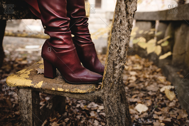 Side view of crop anonymous female in stylish elegant leather boots standing on old stone bench in autumn park