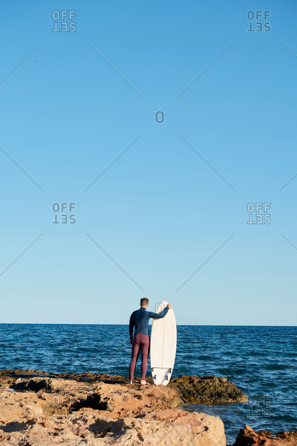 Back view of unrecognizable male surfer in wetsuit standing on rock near sea with surfboard and getting ready for surfing