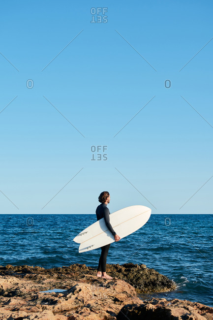 Side view of male surfer in wetsuit standing on rock near sea with surfboard and getting ready for surfing