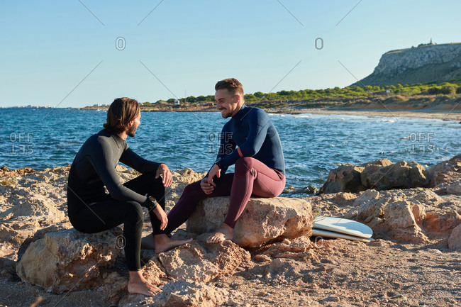 Cheerful male surfers in wetsuits sitting on rocks with surfboards in sea and laughing at jokes together