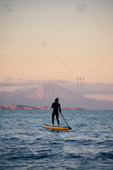 Male surfer in wetsuit rowing on paddle board on sea water on background of mountains at sunset