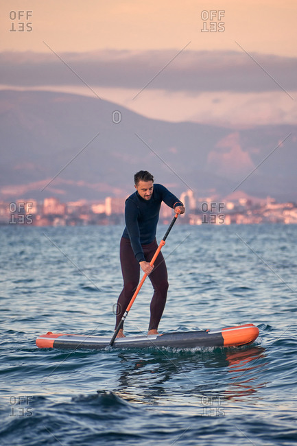 Male surfer in wetsuit rowing on paddle board on sea water on background of mountains at sunset