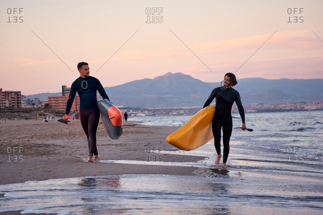 Positive male surfers in wetsuits and with paddleboards walking along wet seashore after surfing at sunset