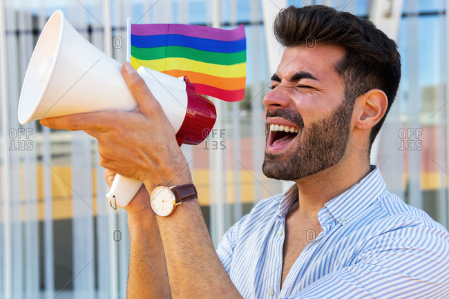 Side view of eccentric gay male with rainbow LGBT flag standing in city and yelling in loudspeaker with closed eyes