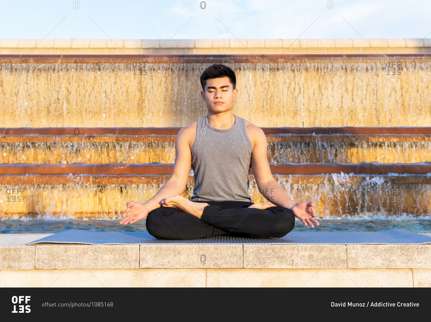 Tranquil focused Asian male in activewear in lotus pose meditating near fountain in city with eyes closed