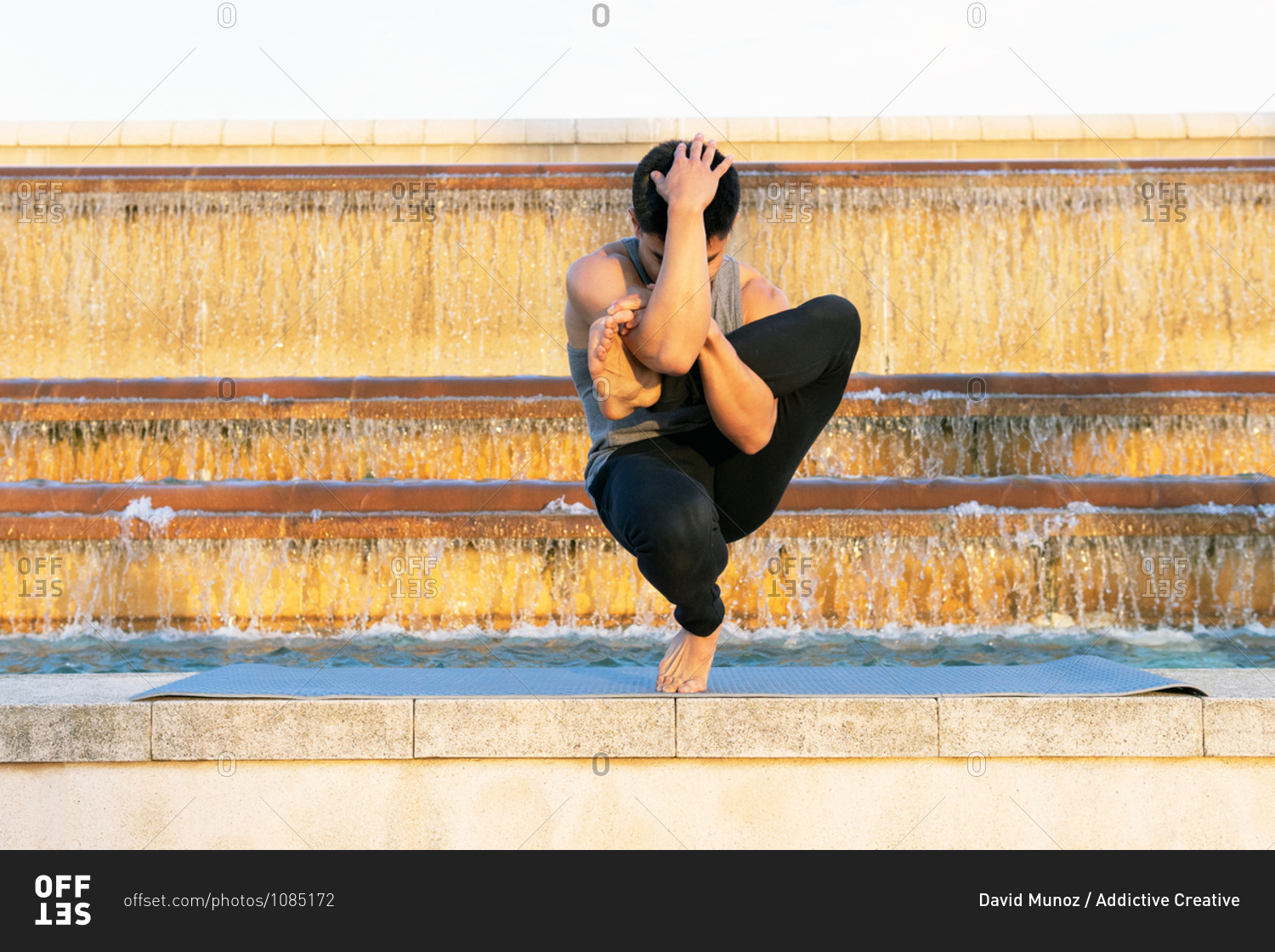 Fit male in activewear doing yoga in Padangusthasana while balancing on leg on mat and practicing meditation near fountain in city
