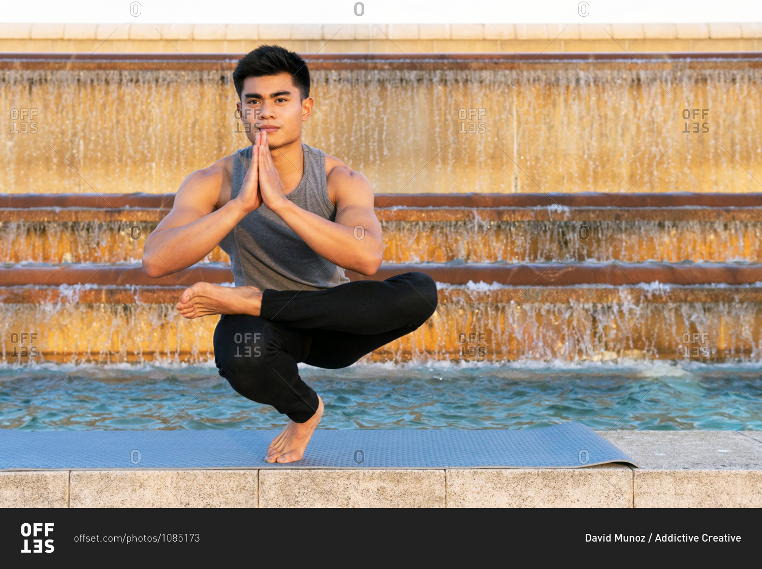 Fit asian male in activewear doing yoga in Padangusthasana while balancing on leg on mat and practicing meditation near fountain in city