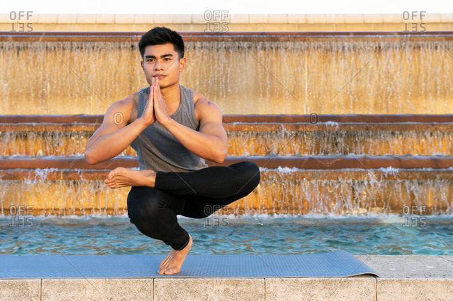 Fit asian male in activewear doing yoga in Padangusthasana while balancing on leg on mat and practicing meditation near fountain in city