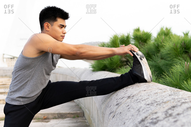 Side view of fit Asian male athlete warming up and stretching legs before workout in city while standing near stone fence and looking away