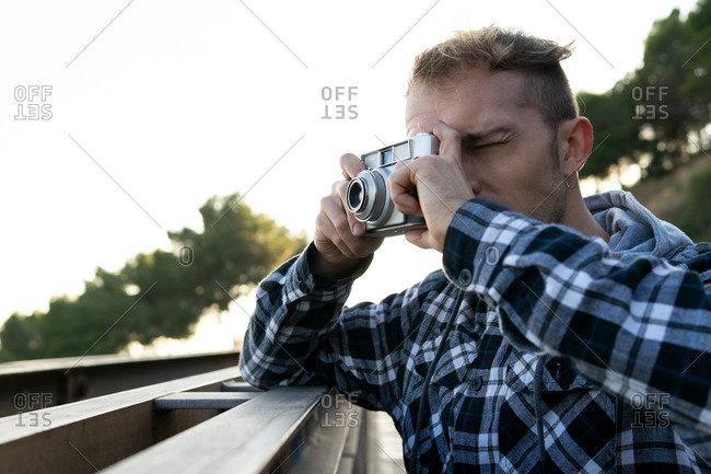 Hipster male photographer in casual checkered shirt standing on bridge and taking pictures of nature with photo camera