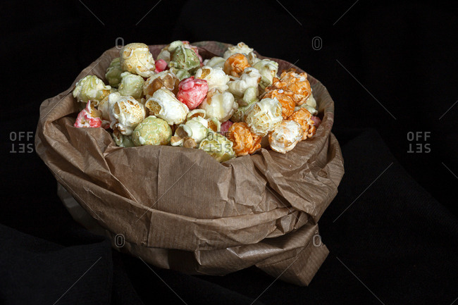 Heap of varicolored sweet popcorn in paper bag placed with yellow corn seeds on black background in studio