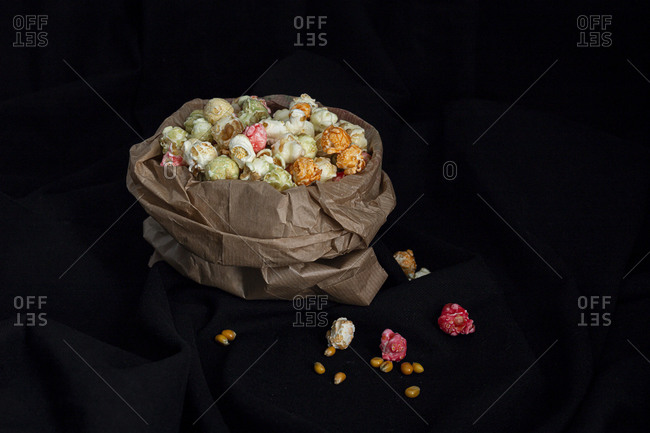 Heap of varicolored sweet popcorn in paper bag placed with yellow corn seeds on black background in studio