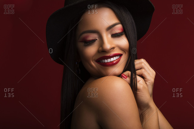 Cheerful female with red lips and in black hat standing on red background and smiling charmingly while touching head and looking away