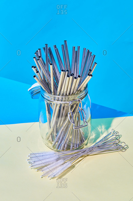From above of many metal reusable straws placed in glass jar on table with cleaning brushes on blue background in studio