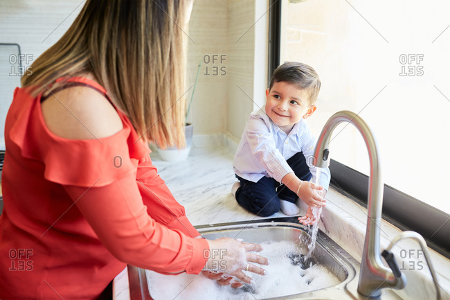 Delighted mother standing at sink in kitchen and washing dishes together with adorable little boy
