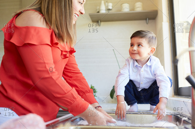 Delighted mother standing at sink in kitchen and washing dishes together with adorable little boy