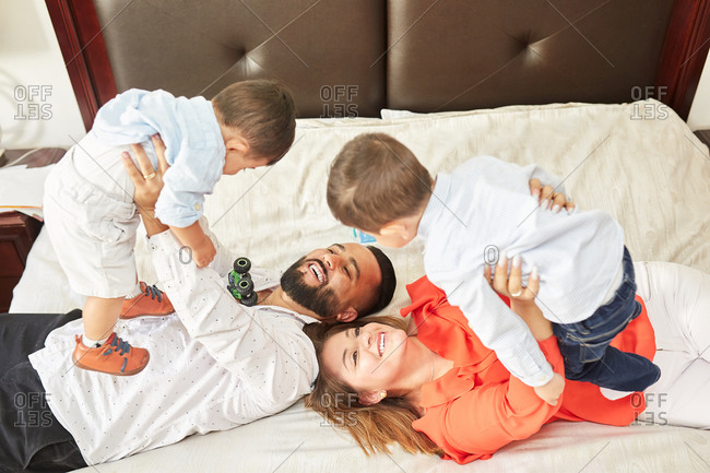 From above of playful Hispanic father tickling adorable laughing children and mother lying on bed and having fun at home
