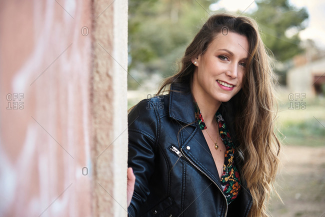 Smiling charming female with long hair and in leather jacket peeping out of wall of shabby building and looking at camera