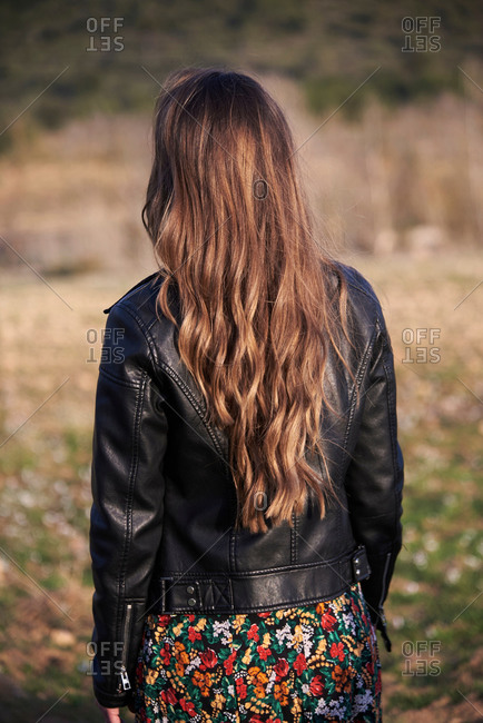 Back view of unrecognizable female in leather jacket and with long wavy hair standing in meadow