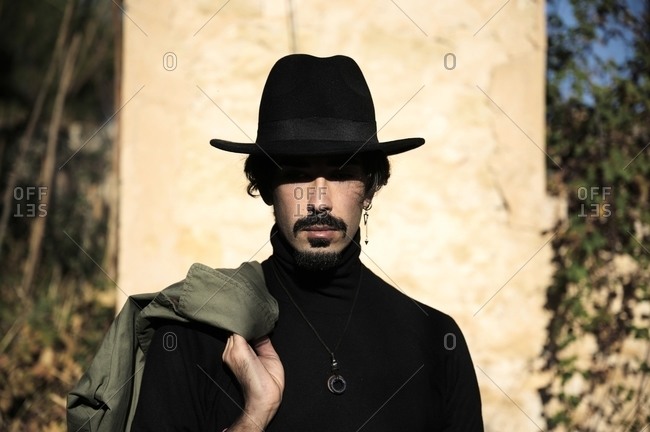 Young bearded guy in stylish black clothes and hat smoking cigarette and looking down thoughtfully while standing near shabby concrete wall on street